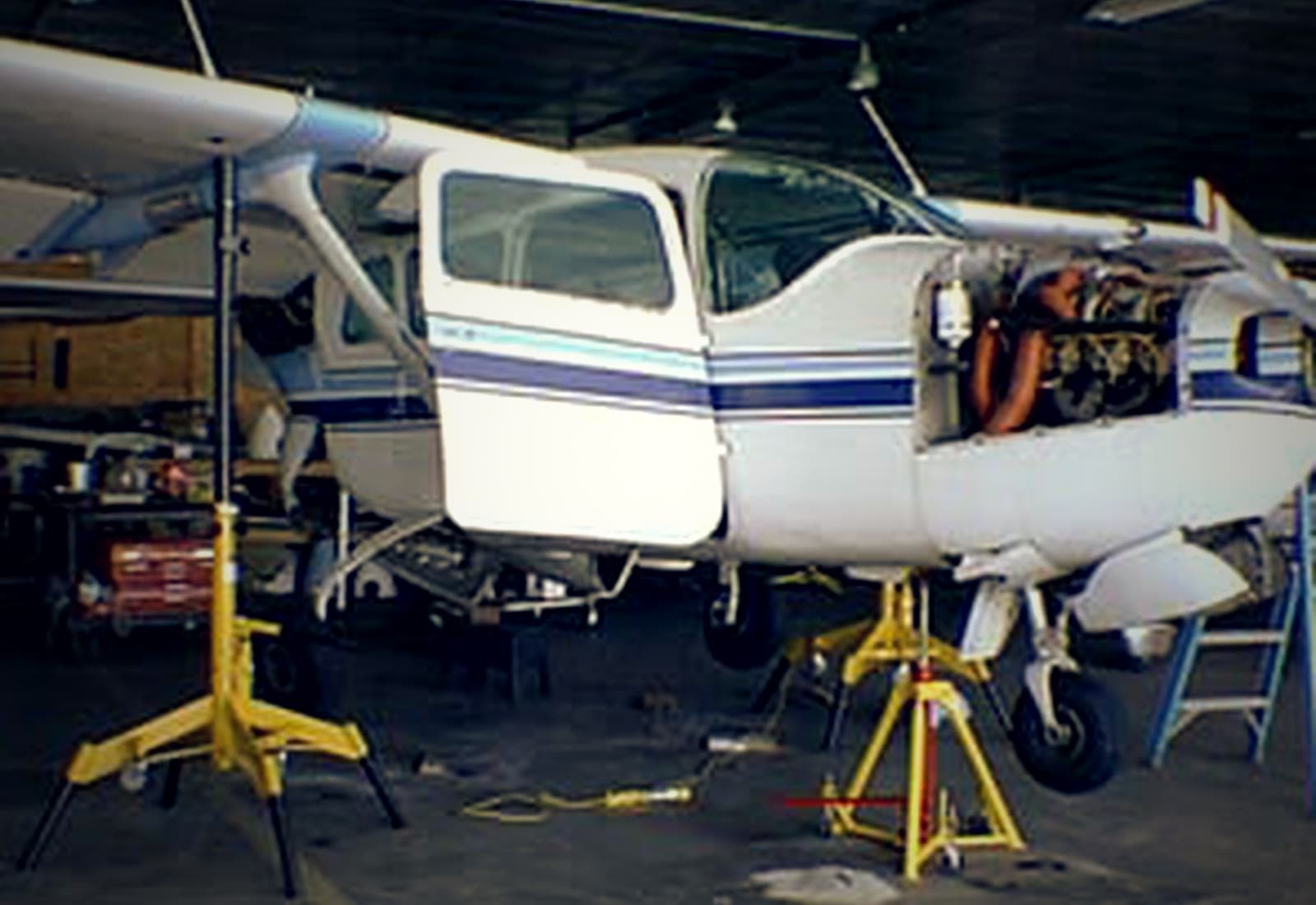 A high-wing Cessna on yellow maintenance jacks in the hangar, cowling removed.